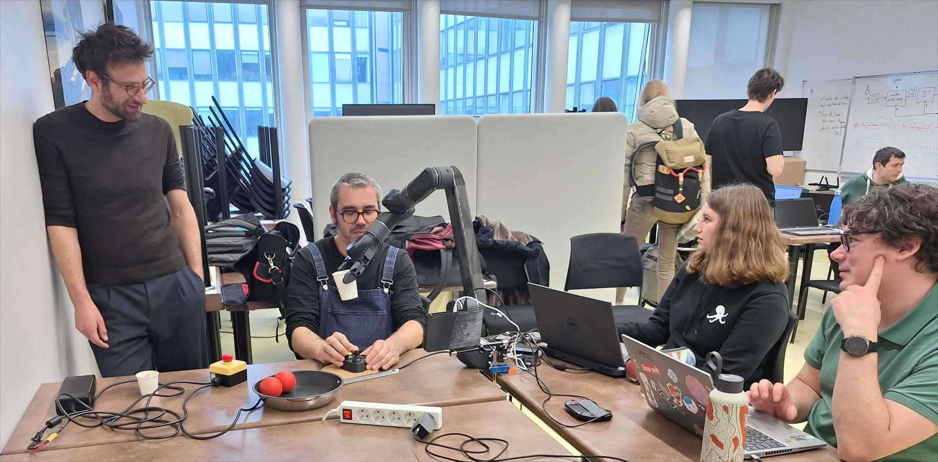 A working session around the assistive arm: an operator drives it with a joystick while engineers and researchers follow on their laptops